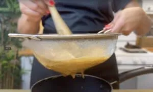 Person straining sauce through a metal sieve into a pot in a kitchen setting.